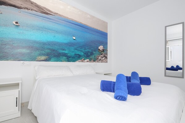 A hotel room with a large bed, white bedding, and a painting of a coastal scene.