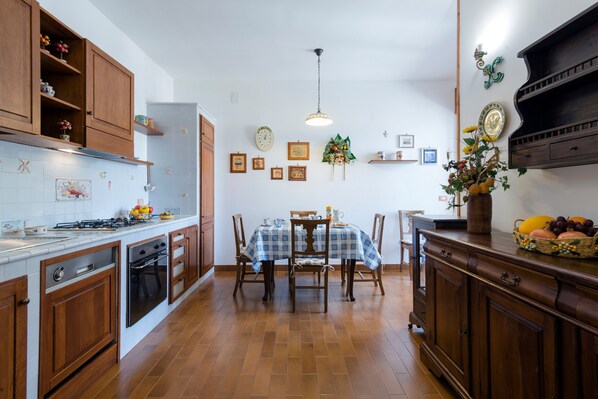 A kitchen with wooden cabinets, a dining table with chairs, a hanging light fixture, and a wall-mounted shelf with decorative items.
