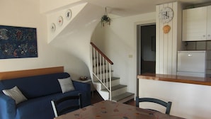 A living room with a blue sofa, a dining table with a fish-patterned tablecloth, a staircase, and a kitchen area with a clock and cabinets. - Apartment "Golfo Stella" With Sea View and Near the Sea (CAPOLIVERI)