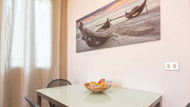 Un coin repas comprenant une table, des chaises, un bol de collations et un tableau représentant un bateau sur une plage, accroché au mur.