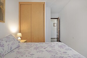 Un dormitorio con cama, una mesita de noche con lámpara, un armario de madera y un pasillo que conduce a un baño.