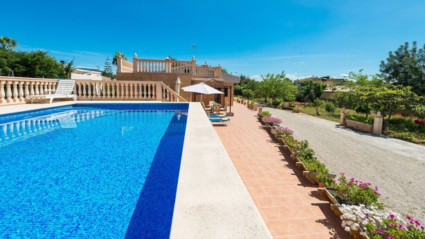 A swimming pool with a clear blue water, a patio area with chairs and a table, and a large building with a balcony in the background.