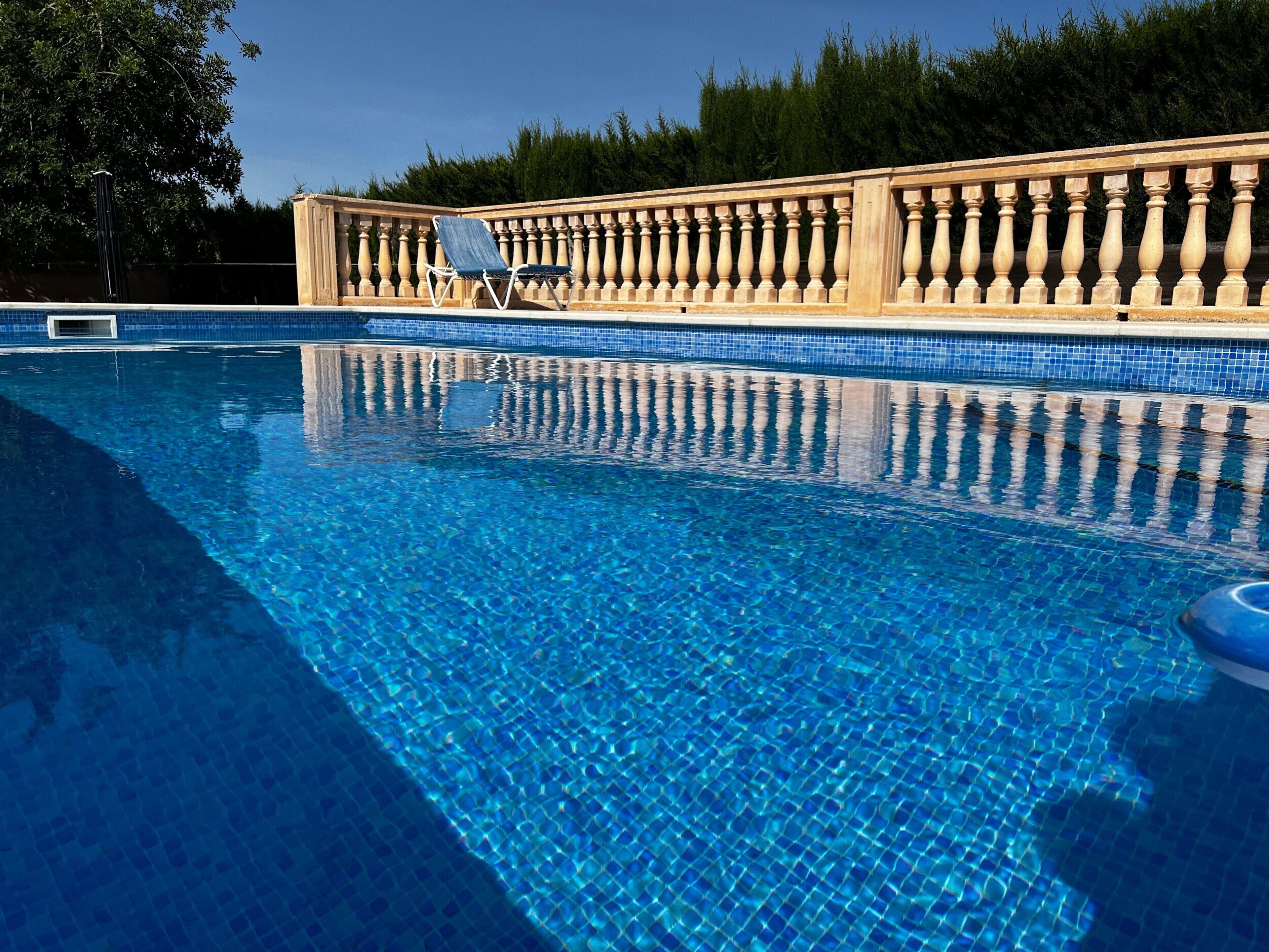 A clear blue swimming pool with a tiled edge, a wooden balustrade, and a single lounge chair.