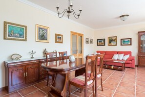 A dining area with a wooden table, chairs, and a red sofa. There are framed pictures on the walls and a chandelier above the table.