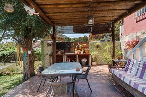 Een overdekt terras buiten met een tafel en stoelen, een bank en een barbecuehoek.