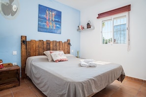 Slaapkamer met een houten hoofdbord, een bed met een grijze spreidek, een wandventilator, een schilderij, een raam met metalen tralies en een plank met boeken en een mand.
