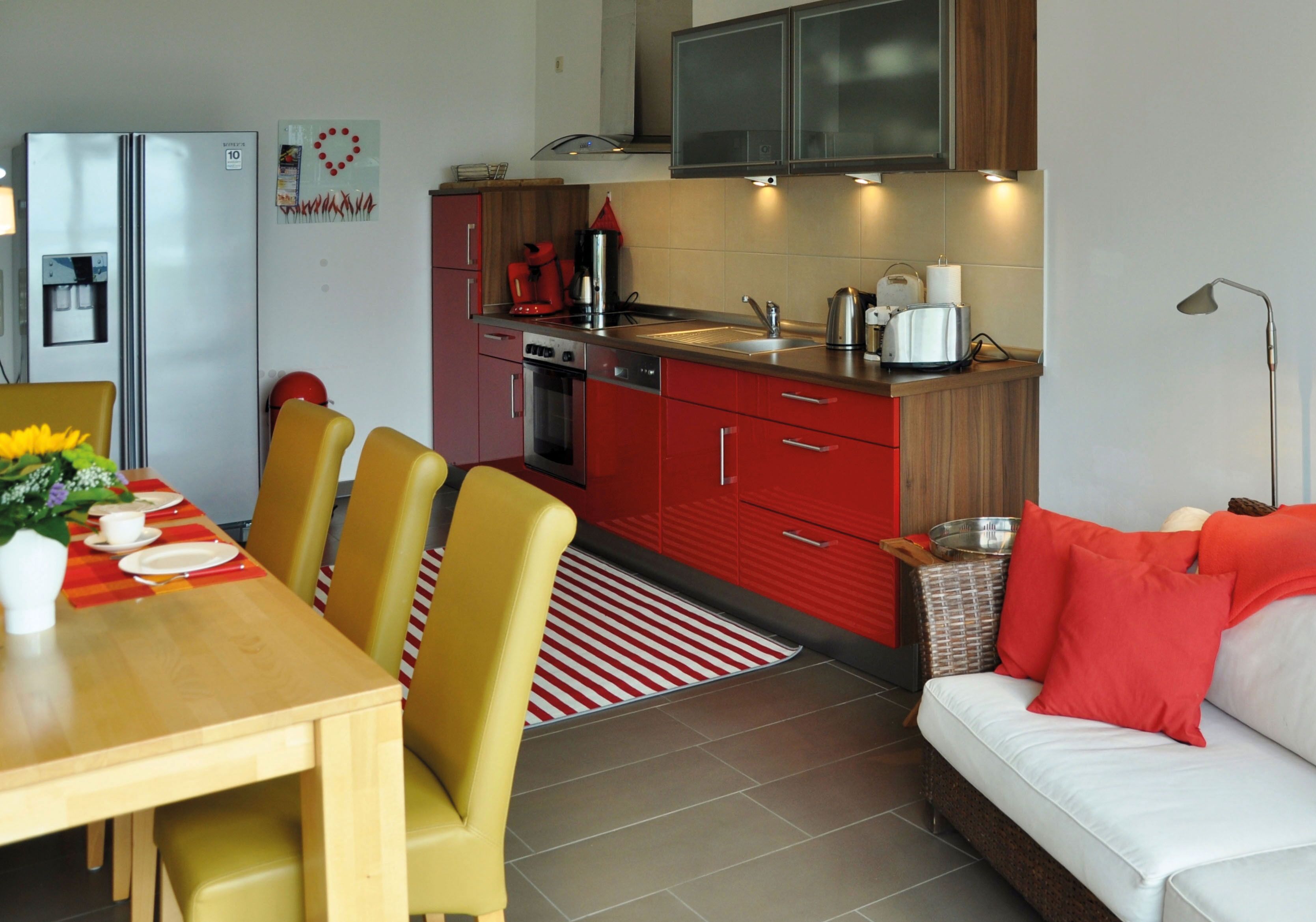 A compact kitchen with a refrigerator, stove, sink, and red cabinets. There is a dining table with chairs and a small sofa with red cushions.