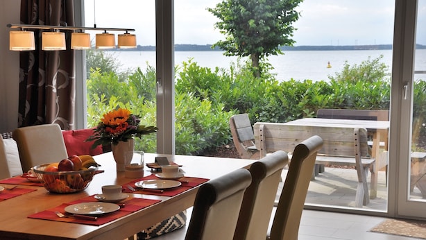 A dining area with a table set for two, a vase with flowers, and a view of the water. - Beach house, 200 sqm. Wf., Directly at the Plauer See, Müritz, 30m to the beach (Fünfseen Lenz Süd)