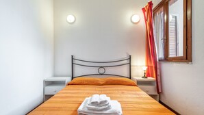 Een keurig opgemaakt bed met een gestreept laken, een klein nachtkastje met een lamp, en een raam met gordijnen.