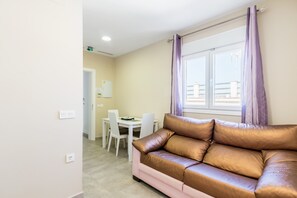 A living room with a brown leather sofa, a dining table with chairs, a window with purple curtains, and a white wall with a light switch.