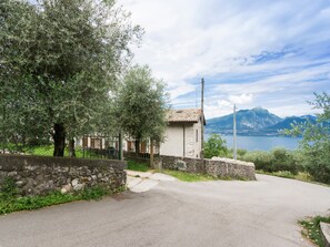 Une route de campagne mène à un bâtiment en pierre surmonté d’un toit de tuiles, entouré d’oliviers et offrant une vue sur des montagnes et un lac.