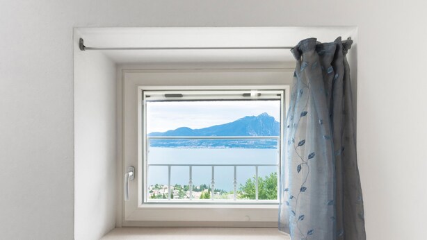 Ein Fenster mit Blick auf einen See und Berge, ein weißer Vorhang und eine weiße Wand.