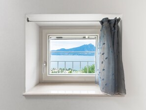 Une fenêtre donnant sur un lac et des montagnes, un rideau blanc et un mur blanc.