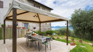 Eine überdachte Terrasse im Freien mit Tisch und Stühlen, umgeben von Topfpflanzen und Blick auf einen See und Berge.