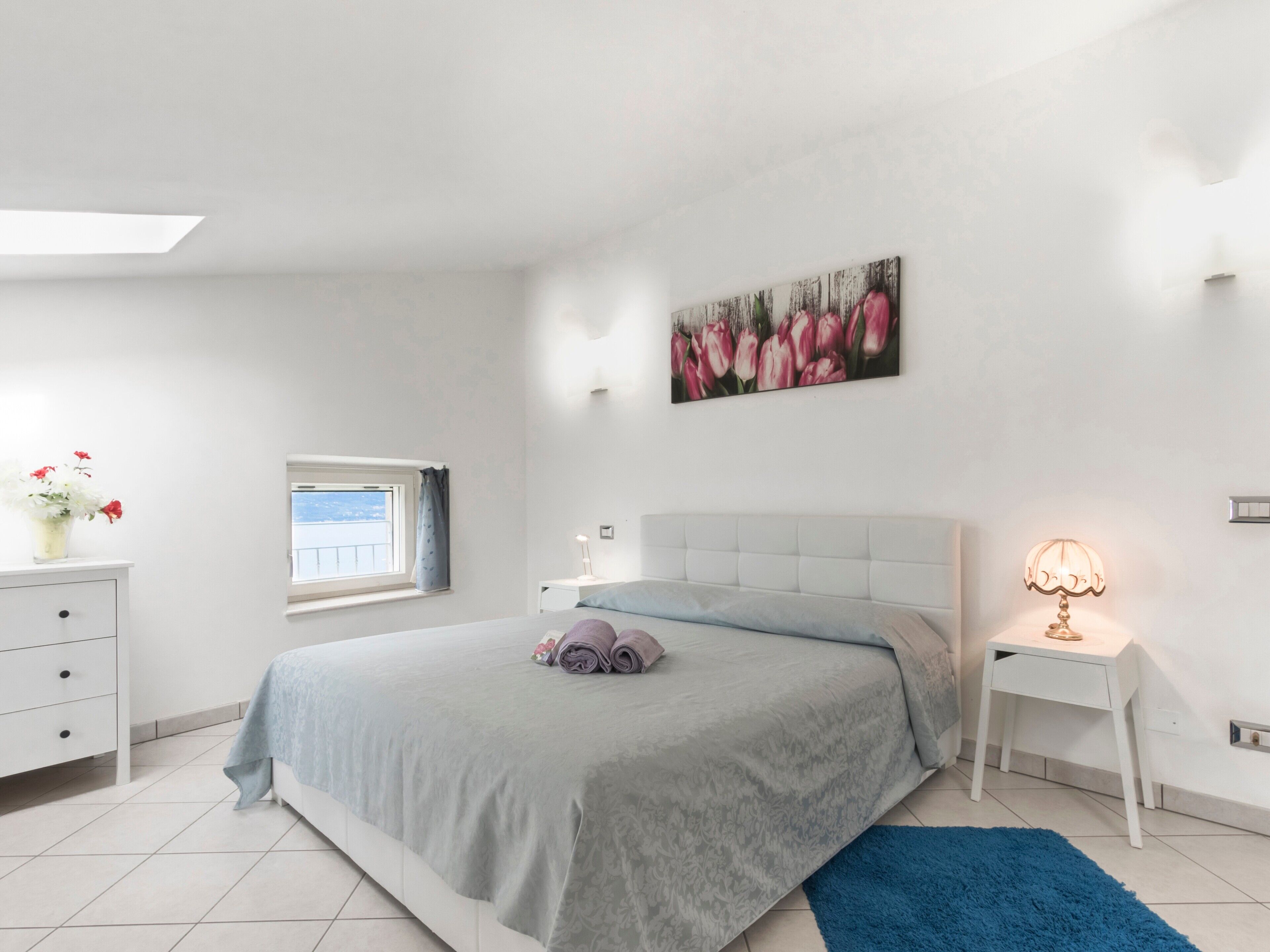 Een slaapkamer met een bed, nachtkastje, lamp, raam en een schilderij.