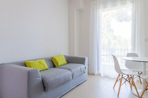 A modern living room with a grey sofa, two green cushions, a white table with chairs, and a large window with curtains.
