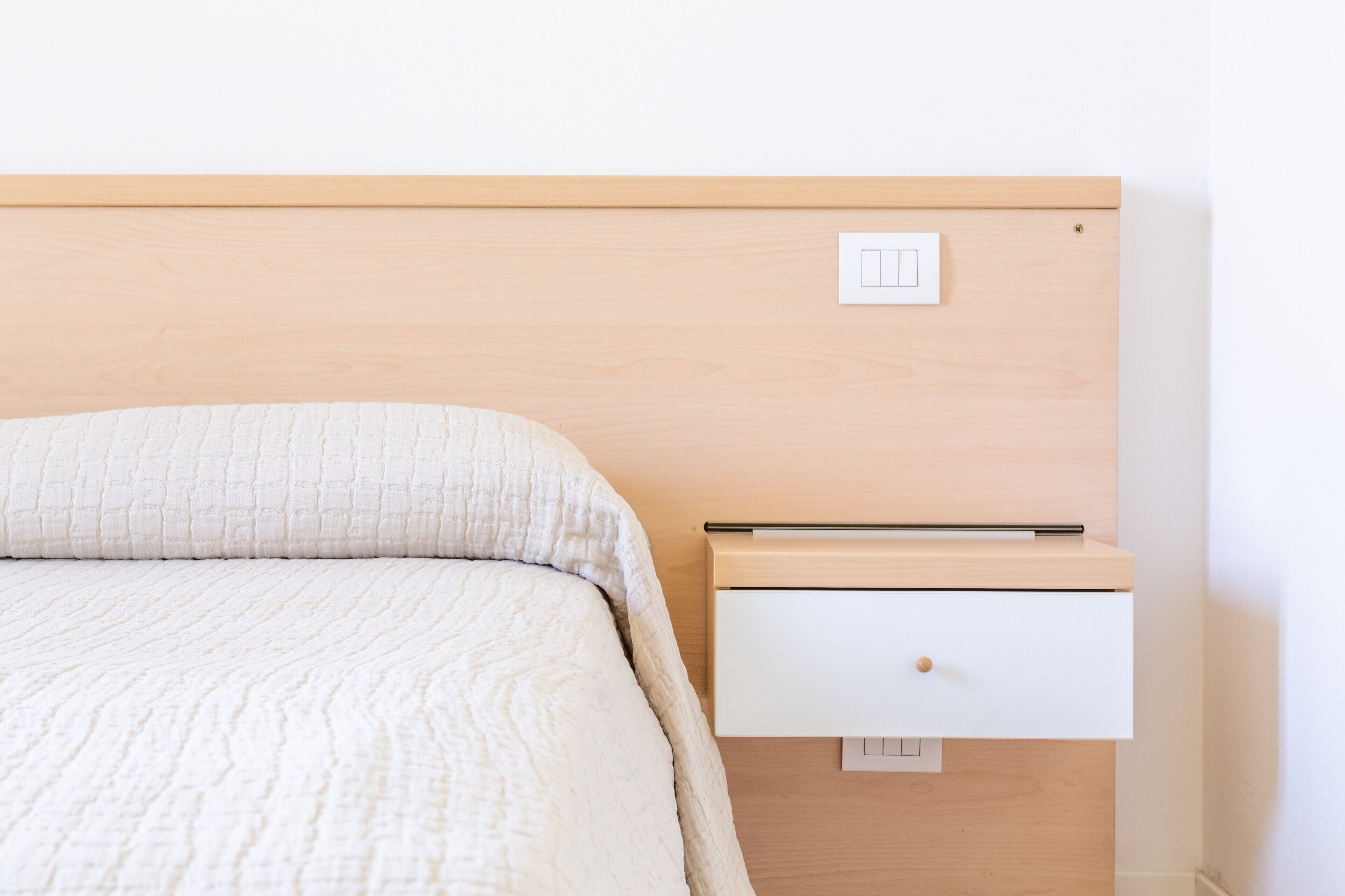 Ein Bett mit strukturierter weißer Decke, einem Holzkopfstück, einer Nachttischschublade und einem Lichtschalter.