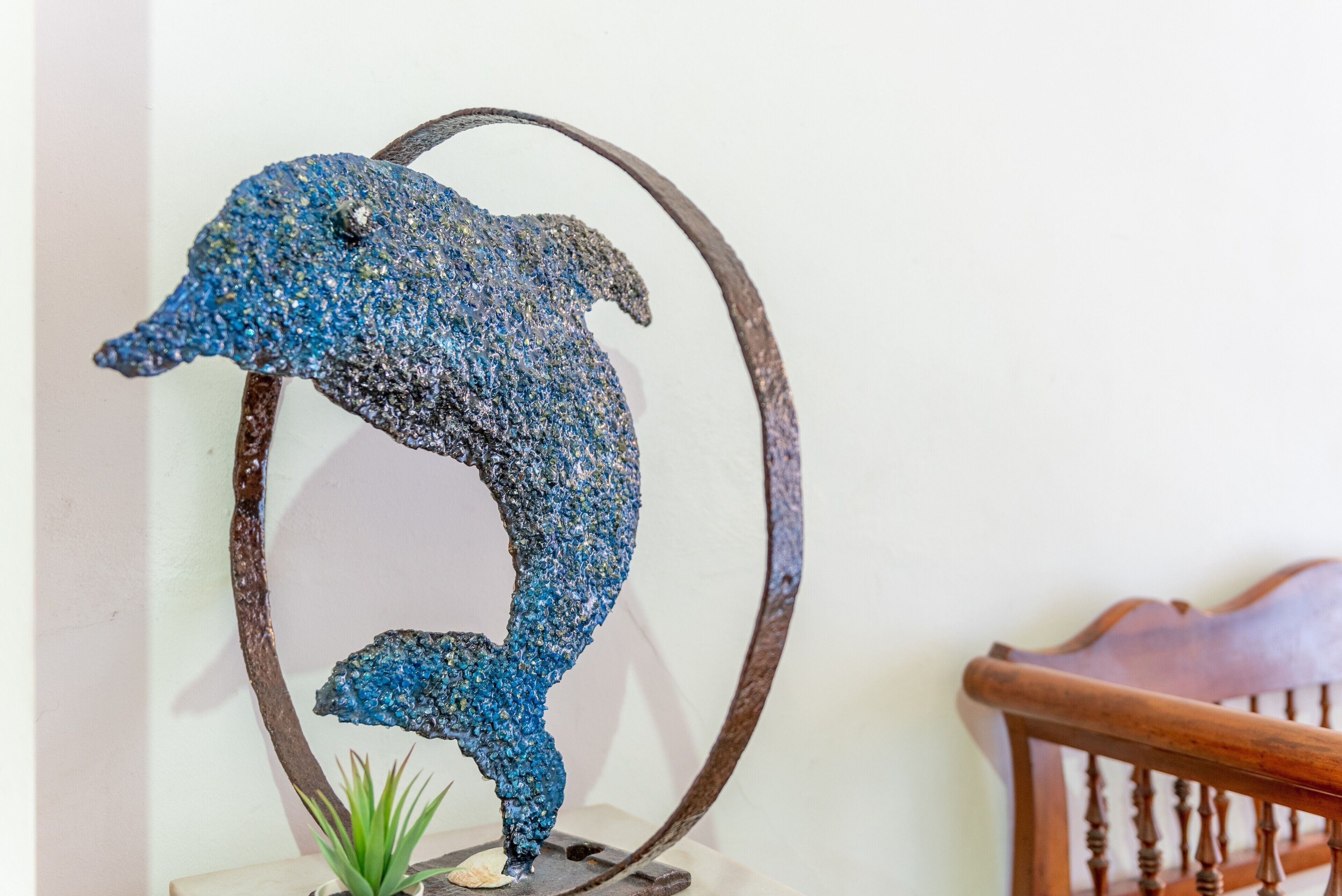 Une sculpture en métal représentant un dauphin, posée sur un socle circulaire, une chaise en bois et une petite plante au premier plan.