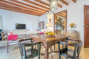 A dining area with a wooden table, chairs, a television, and a kitchen in the background.