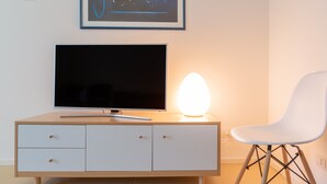 A modern room with a flat-screen TV on a wooden cabinet, a white chair, and a wall-mounted light. - Modern and with Pool - Apartment Baseventuno 1 (Sirmione)