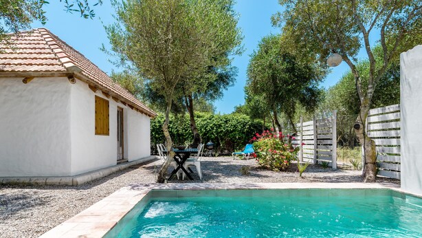 Uma piscina com deck ladrilhado, um edifício branco com telhado ladrilhado e um jardim cercado com árvores e arbustos.