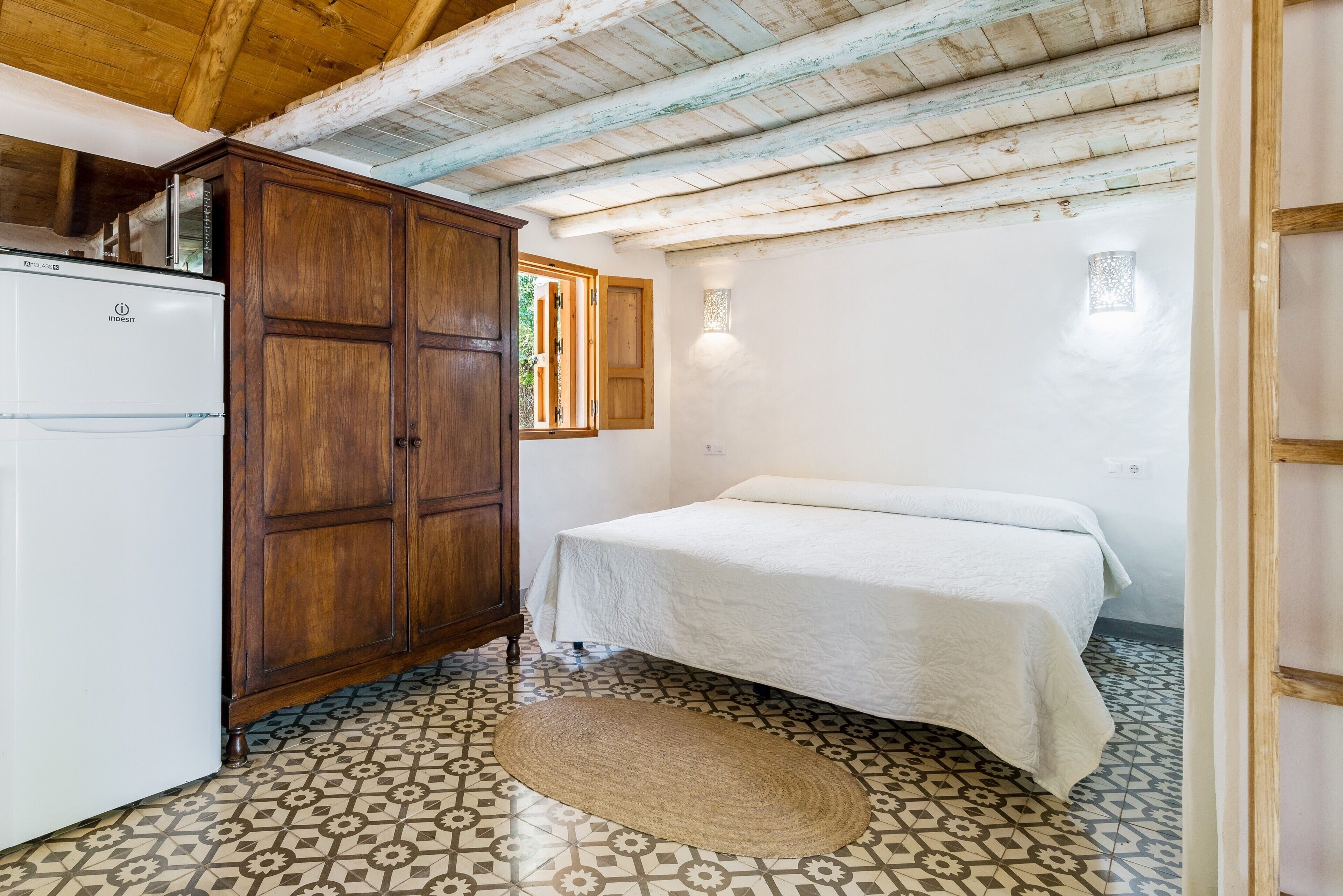 Quarto com cama, guarda-roupa de madeira, frigorífico branco, chão estampado e janela com vista.