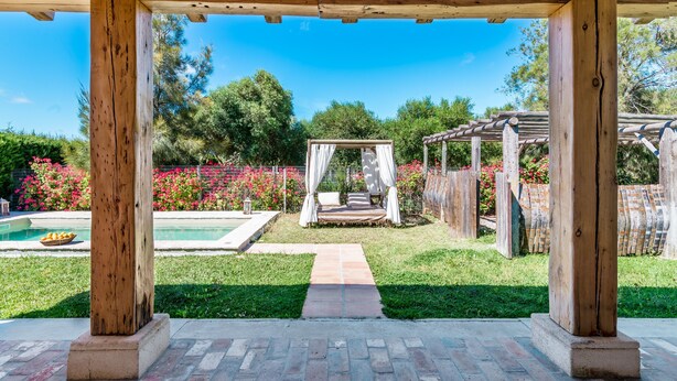 Een houten pergola met een overdekte zithoek, een pad van baksteen en een zwembad op de achtergrond.