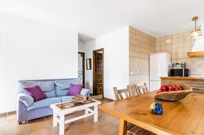 Una sala de estar con un sofá, una mesa de centro, una mesa de comedor con sillas, una zona de cocina con microondas y un frutero.