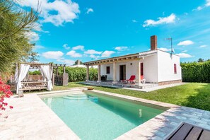 Un área junto a la piscina con un pequeño edificio, una pérgola con cortinas blancas y un cielo azul despejado.