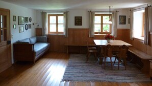 Ein gemütliches Zimmer mit Holzboden, einer Couch, einem Esstisch mit Stühlen und zwei Fenstern mit Vorhängen.