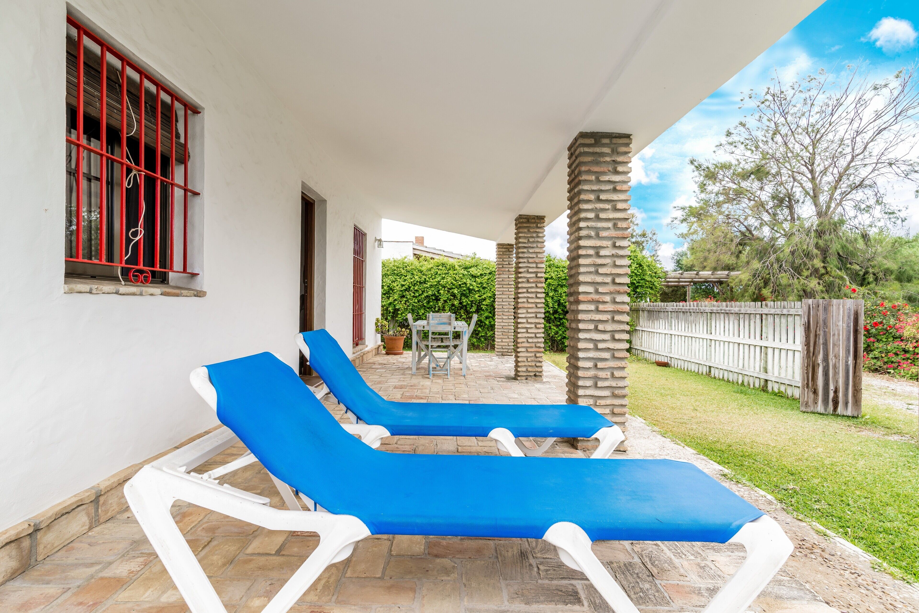 Eine überdachte Terrasse mit zwei blauen Liegestühlen, einer Steinpfeiler und Blick auf einen Garten mit einem Holzzaun.