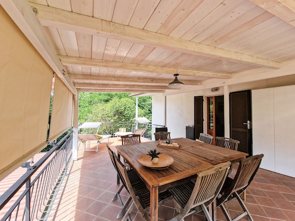 A covered patio with a wooden table and chairs, a ceiling fan, and a view of greenery.