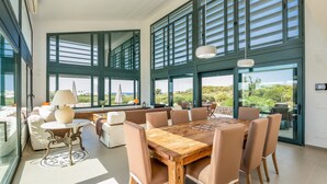 Un comedor amplio con una mesa y sillas de madera, ventanas grandes con persianas y vistas a zonas verdes.