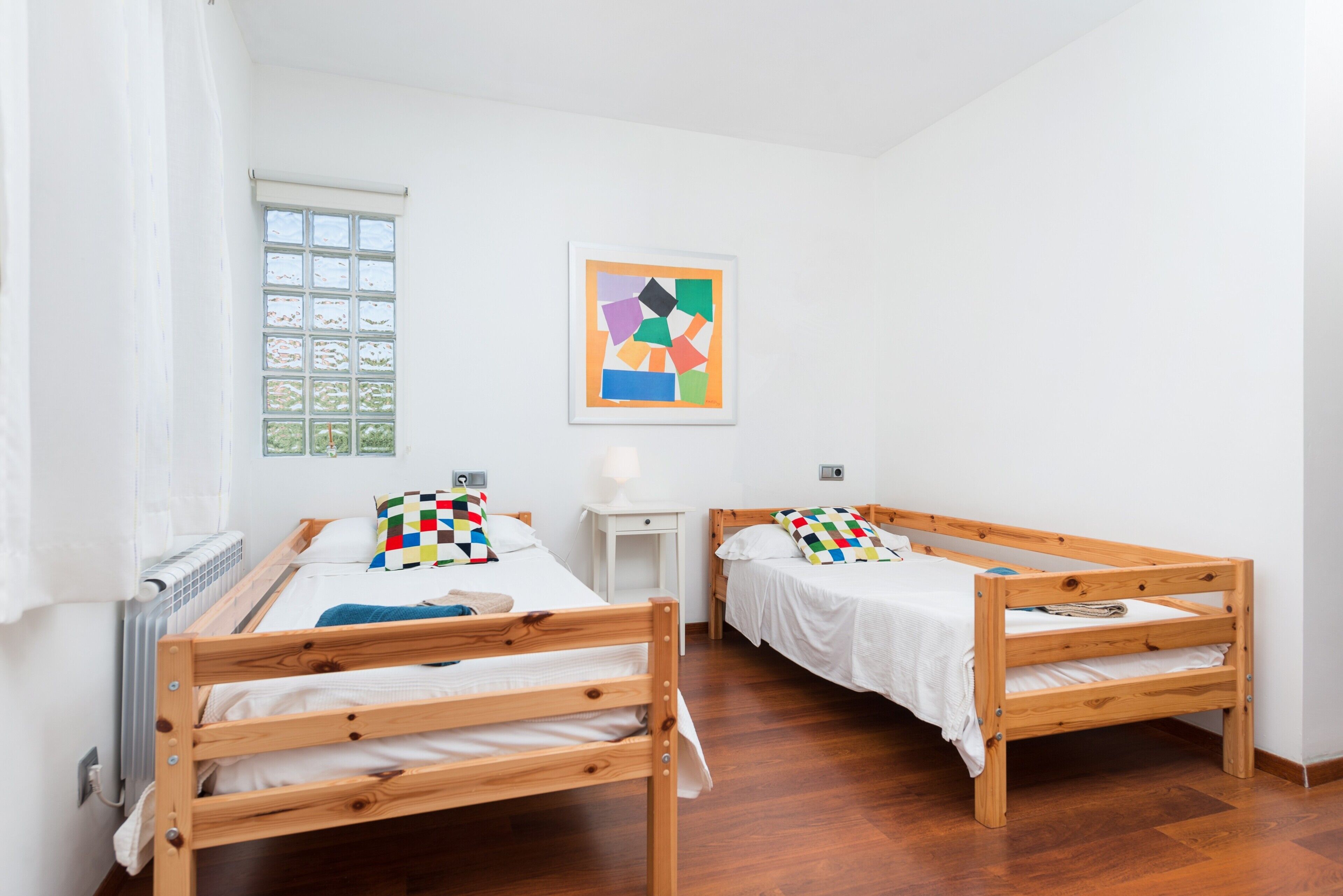 A room with two wooden bunk beds, a window with a view, a radiator, and a framed abstract painting on the wall.