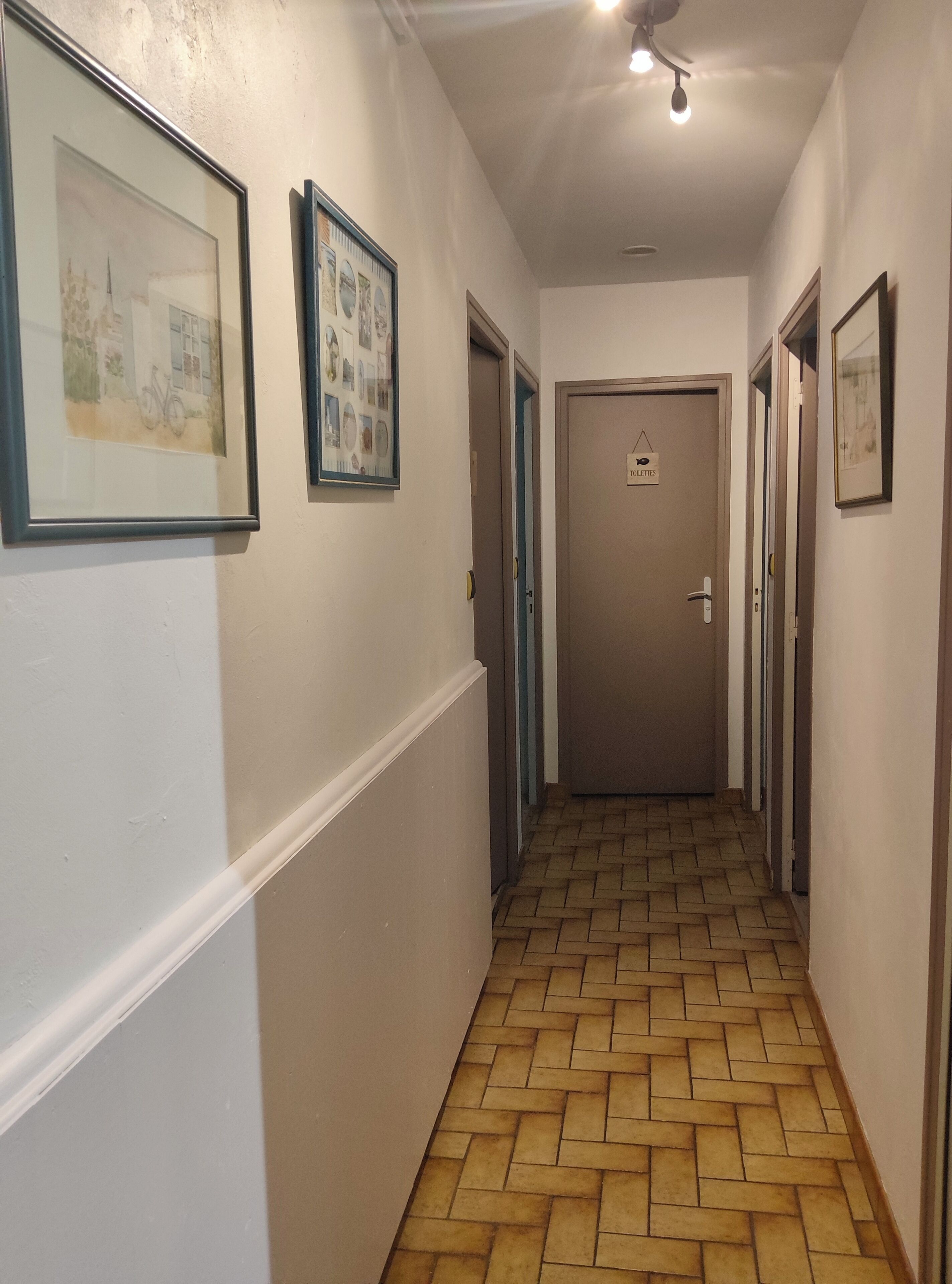 A hallway with framed pictures on the walls, a tiled floor, and a door labeled "TOILETIES".