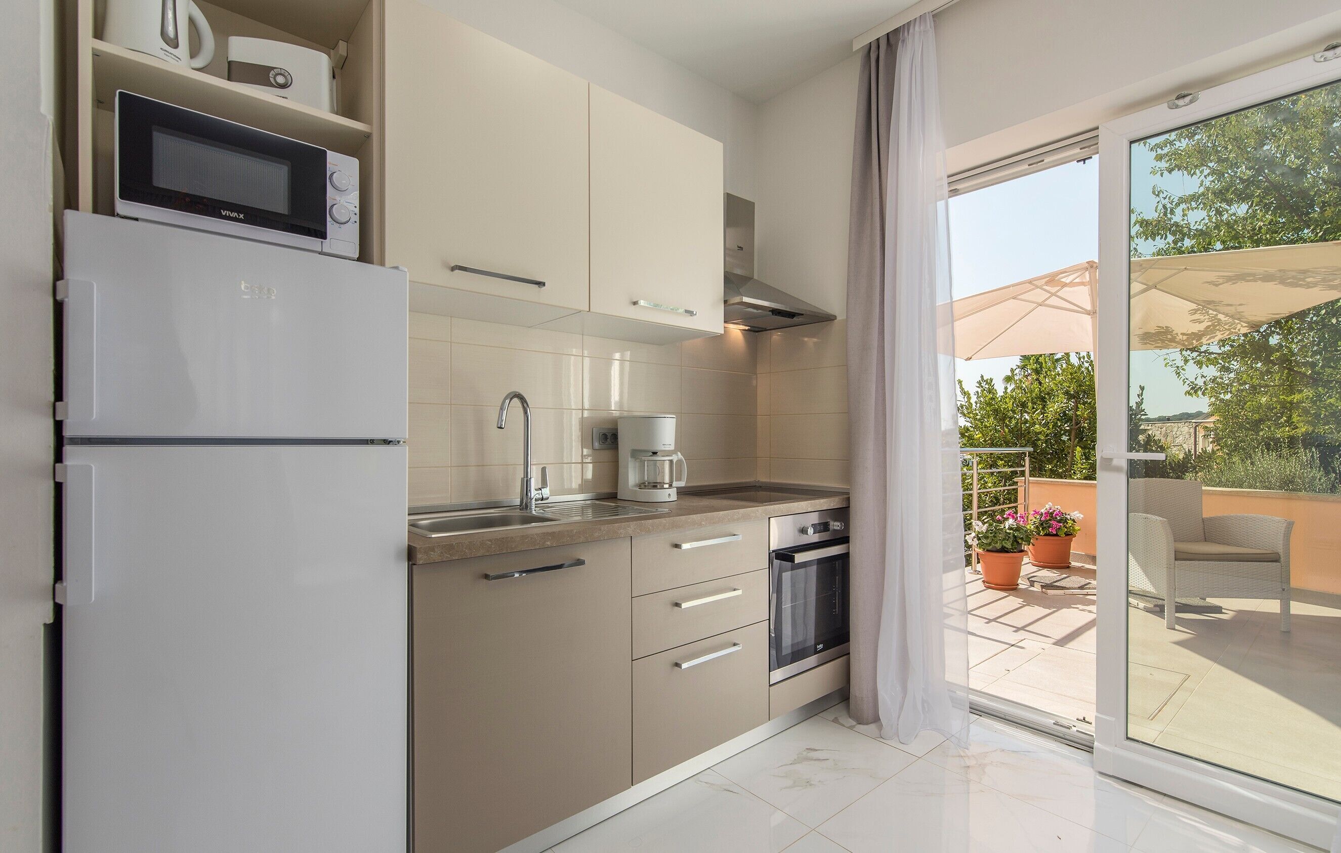 Eine moderne Küche mit Mikrowelle, Kaffeemaschine, Backofen und Glastüren zu einem Balkon mit Blumentöpfen und Gartenmöbeln.