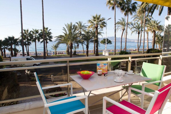 A balcony with a table and chairs overlooking a beach with palm trees and a view of the sea.