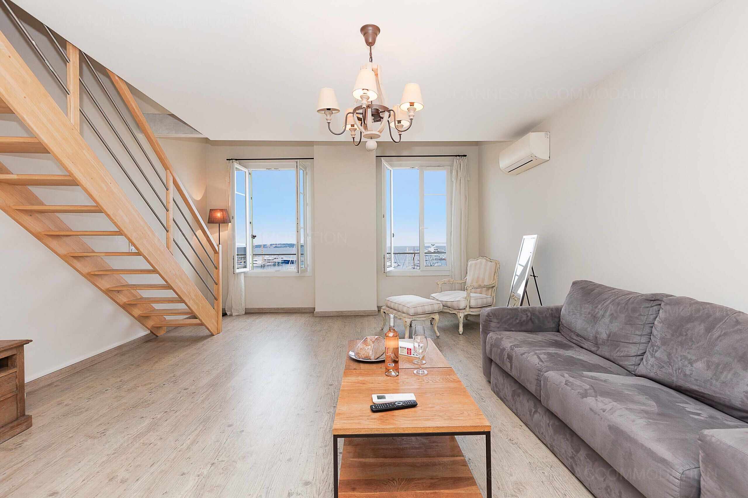 A spacious living room with a wooden staircase, a grey sofa, a wooden coffee table, and a chandelier.