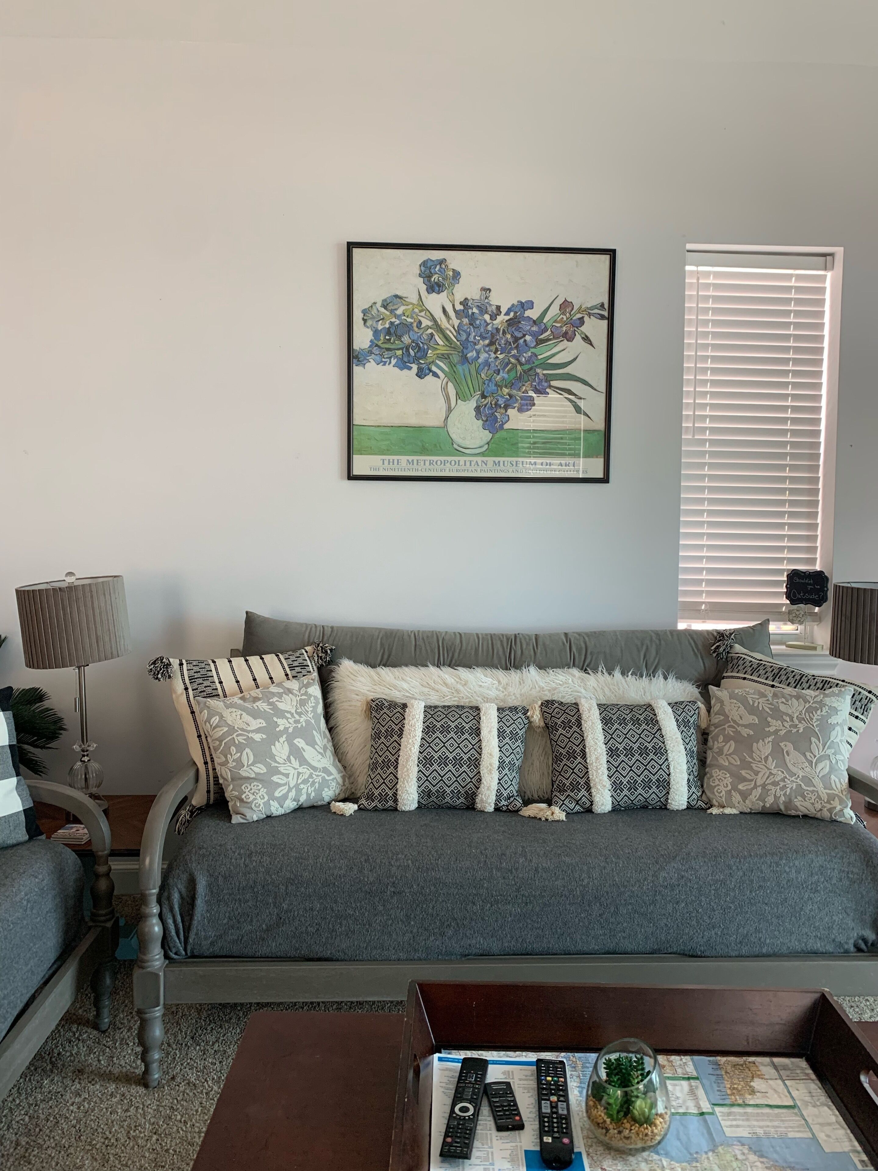 A living room with a grey sofa, patterned cushions, a framed painting, and a coffee table with remote controls and a decorative glass.