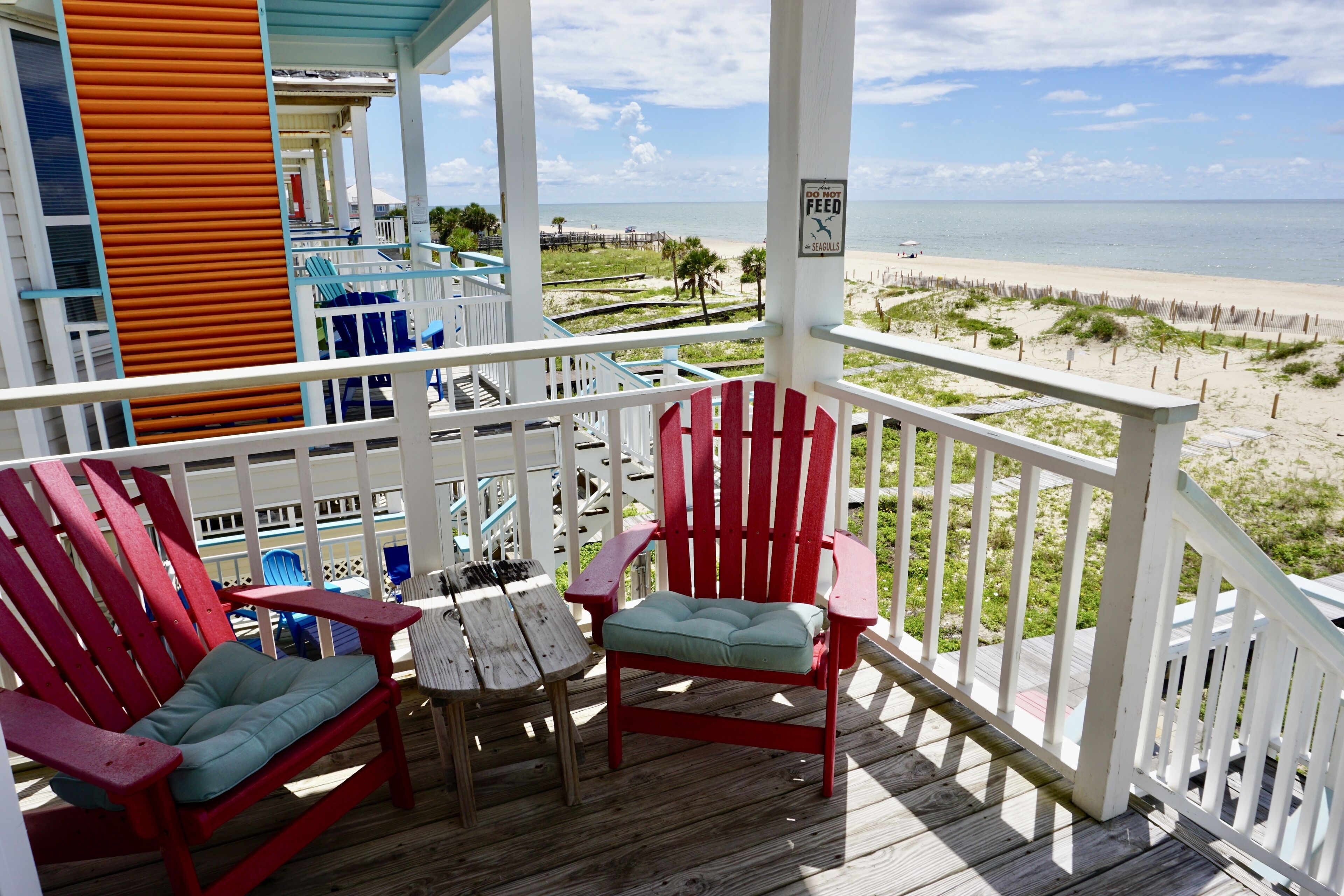A wooden deck with red Adirondack chairs and a table, overlooking a beach with a fence and a sign warning against feeding seagulls.