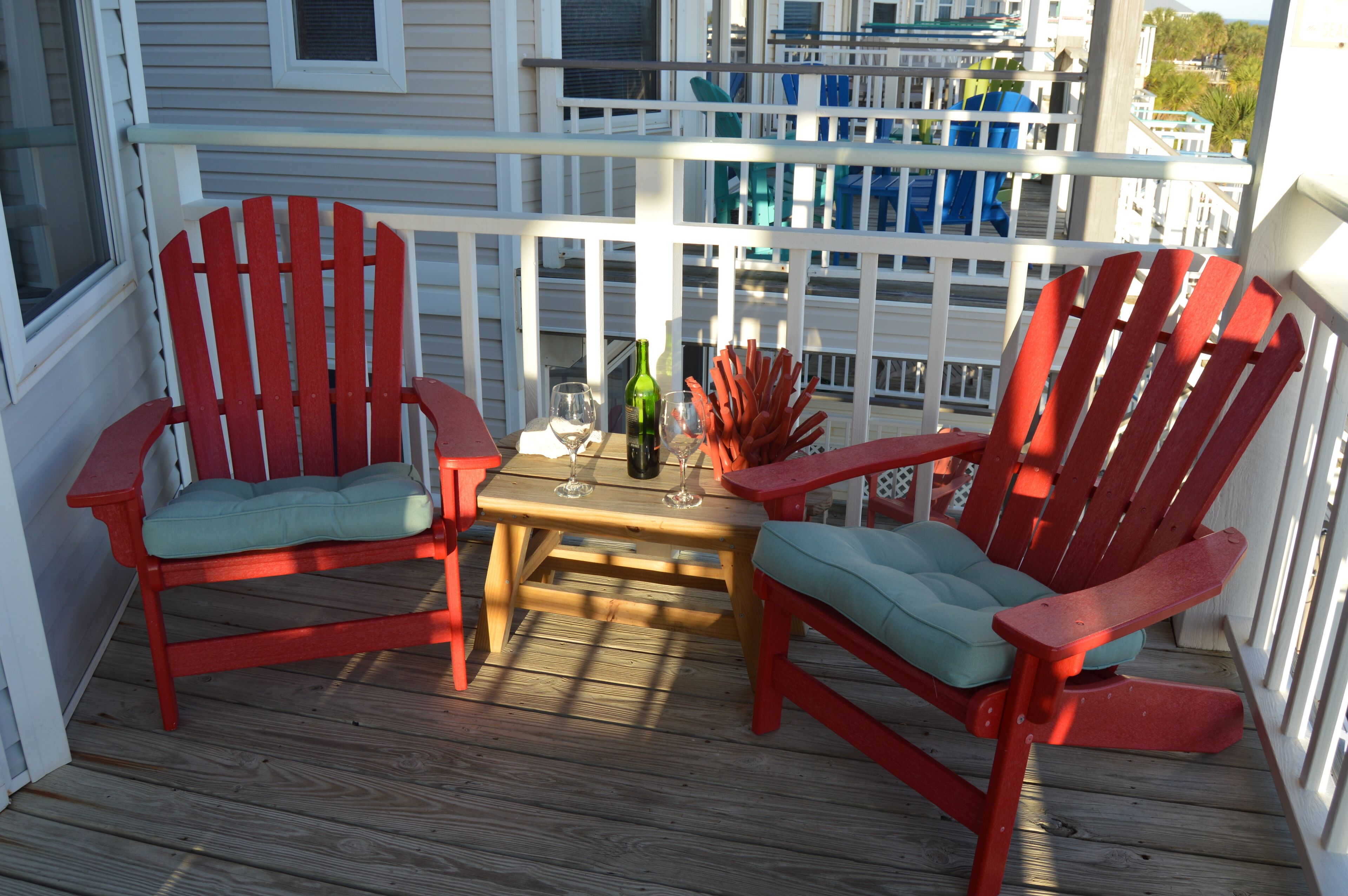 A wooden deck with two red Adirondack chairs and a small table with a bottle and glasses.