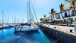 A marina with multiple sailboats docked, a waterfront promenade with palm trees, and buildings with traditional architecture. - Pensión Playa Mogán (Mogan)