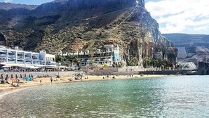 A coastal area with clear waters, a sandy beach, and a rocky cliff with buildings. - Pensión Playa Mogán (Mogan)