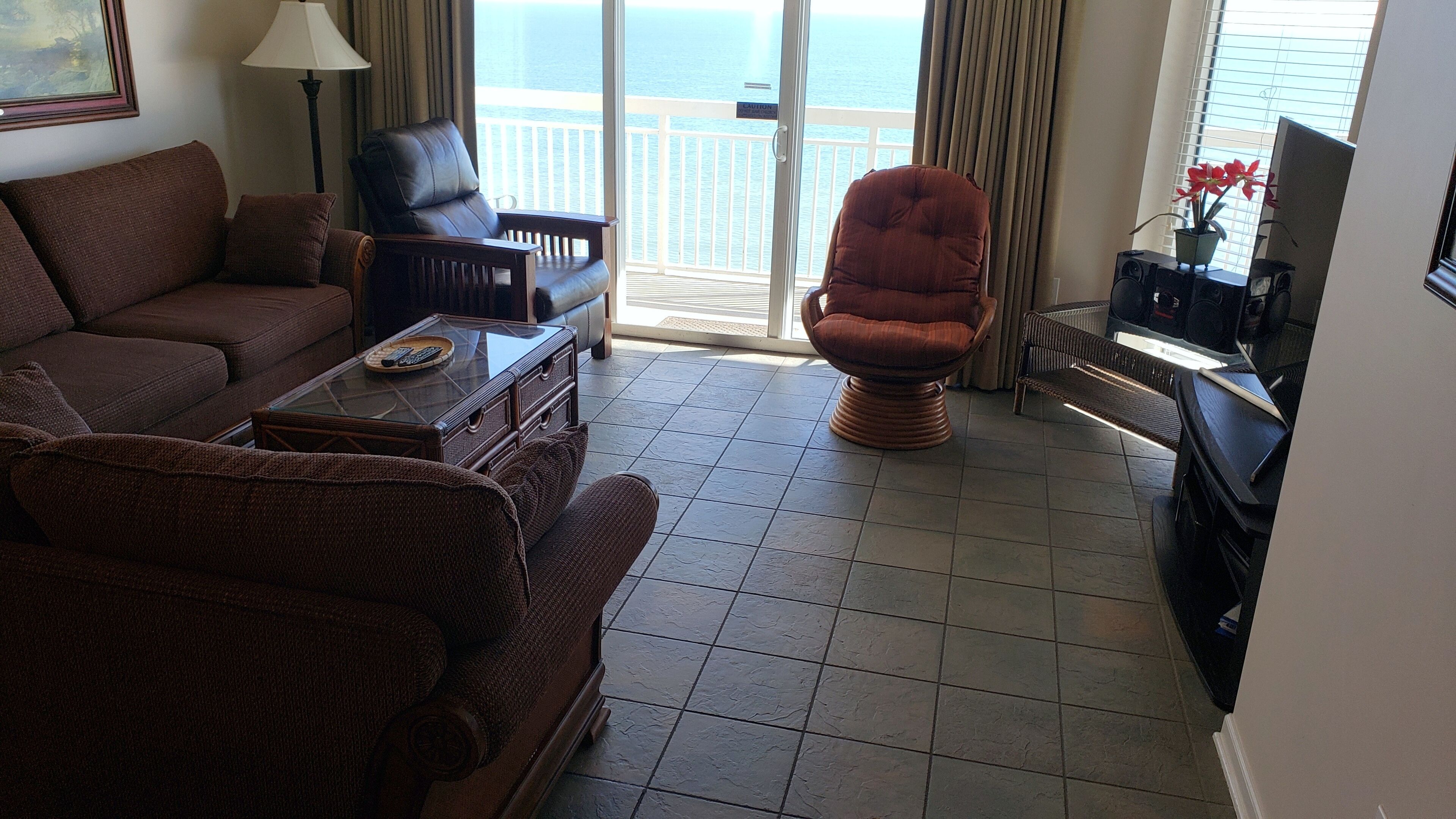 A living room with a sofa, armchairs, a coffee table, and a TV. The room has a balcony with a view of the sea.