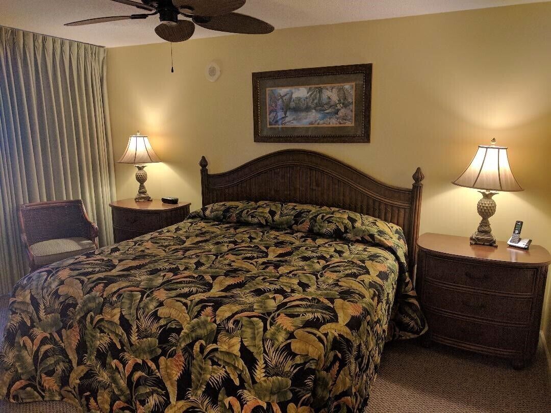 Habitación de hotel con una cama grande, dos mesitas de noche con lámparas, un ventilador de techo, un cuadro en la pared y una ventana con cortinas.