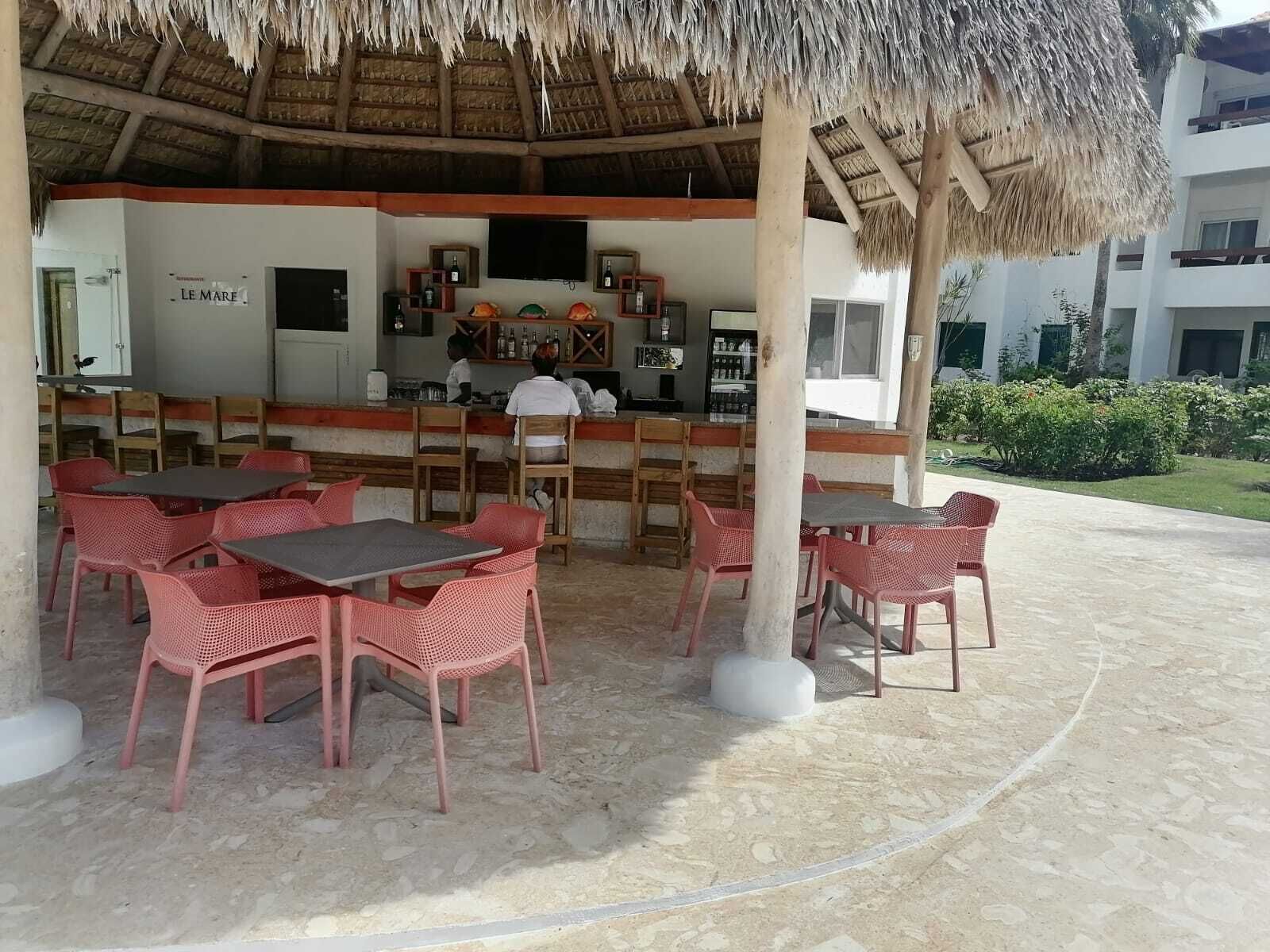 Un espace bar extérieur avec des tables et des chaises, un toit de chaume et une vue sur un bâtiment en arrière-plan.