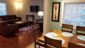 A living room with a sofa, coffee table, and TV, a dining area with a table and chairs, and a wall-mounted TV. - The Blackberry Inn Guest House (Watkins Glen)