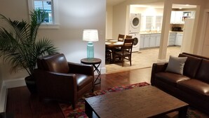 A living room with a brown leather sofa, a wooden coffee table, and a colorful rug. There is a dining area with a table and chairs, and a kitchen in the background. - The Blackberry Inn Guest House (Watkins Glen)