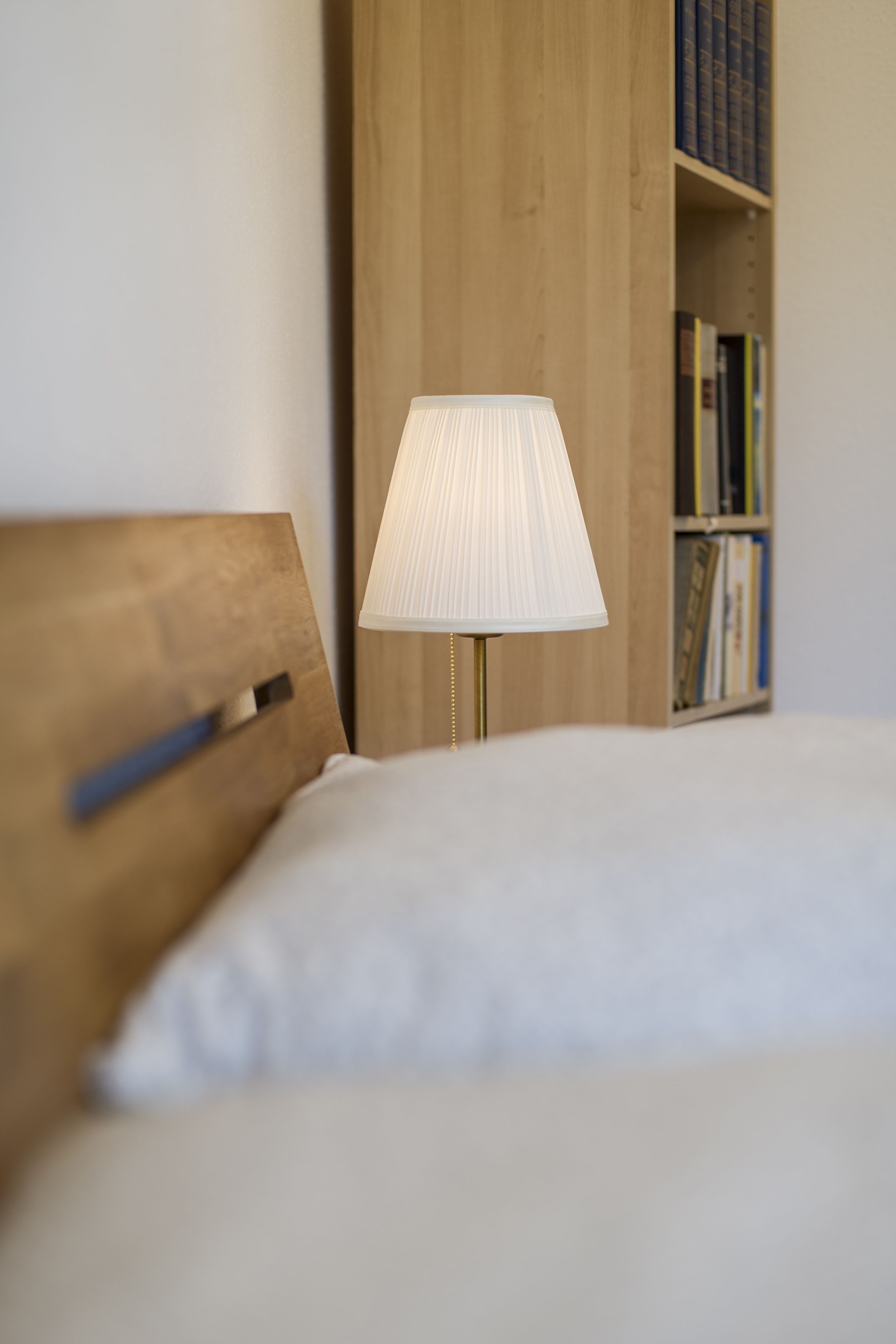 Un letto rifatto con cura, con una testiera in legno, una lampada da comodino e una libreria sullo sfondo.