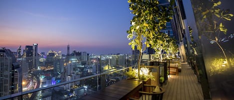 Terraza en la azotea con vista al horizonte urbano al anochecer, que cuenta con cubierta de madera, zona de estar y barandilla de vidrio.